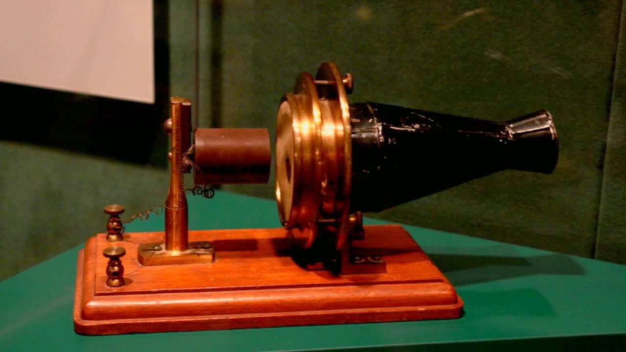 An antique mechanical device with a brass, black, and wooden body, featuring a cone-shaped end, mounted on a polished wooden base against a green background.