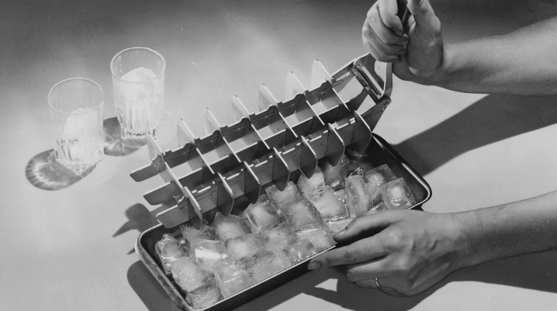 A person pulls the lever of a vintage metal ice cube tray, releasing ice cubes into a tray on a table. Two glasses with ice and water sit nearby.