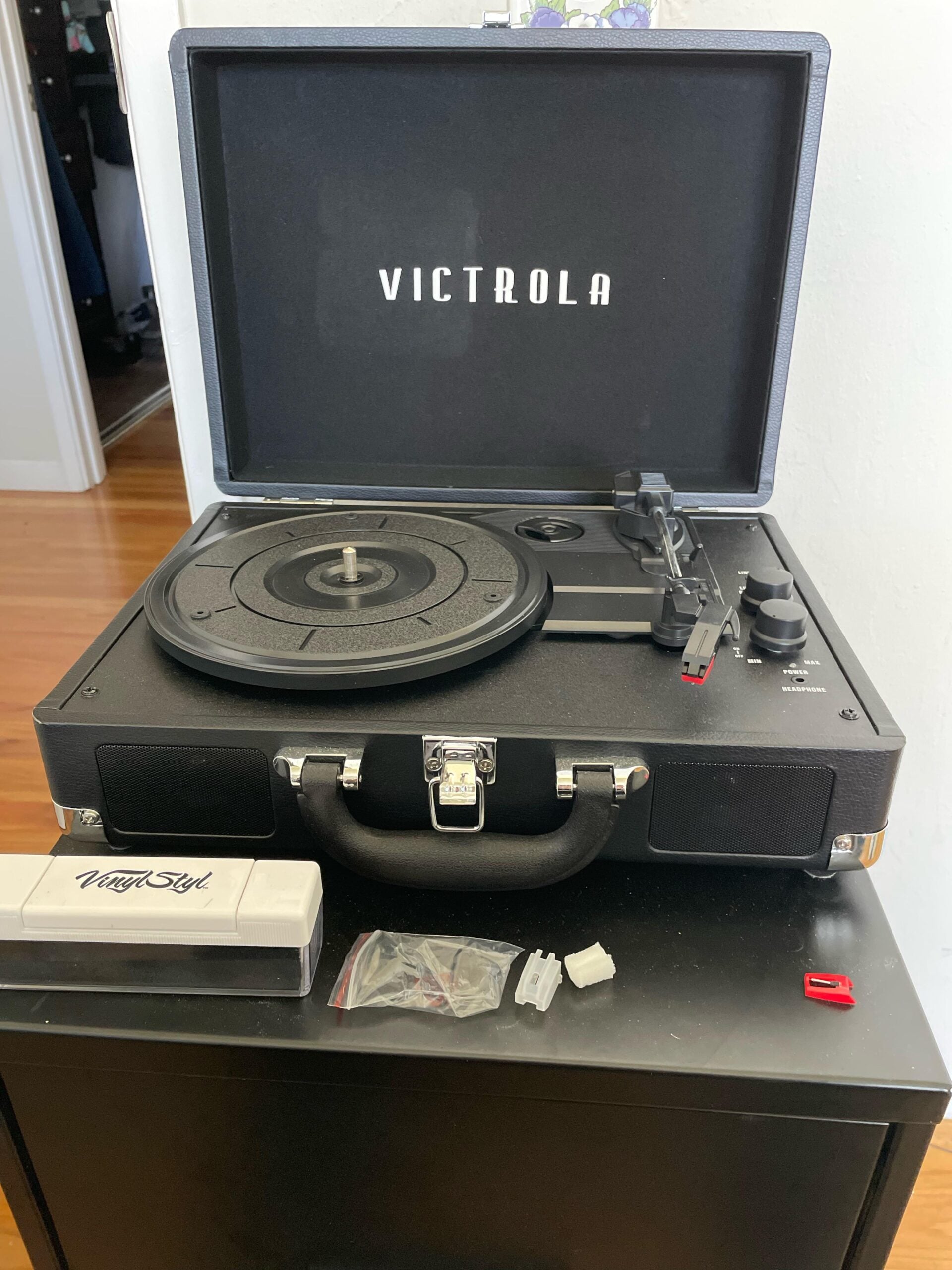 A black Victrola suitcase record player sits open on a small black table, with various small accessories and a white rectangular box labeled “Vinyl Styl” placed in front of it.
