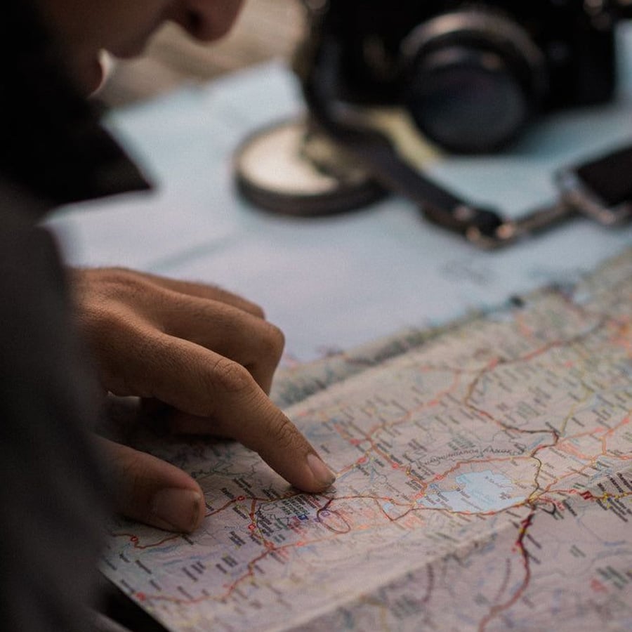 A person points at a location on a detailed map, with a camera, compass, and phone placed nearby, suggesting travel planning or navigation.