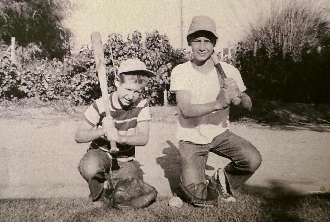 Two boys kneel on grass holding baseball bats, wearing baseball gloves and caps. They smile at the camera, with bushes and trees in the background. The photo is black and white.