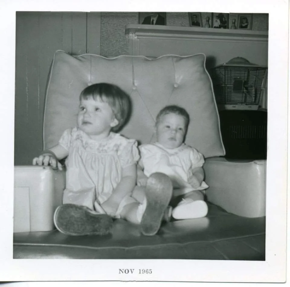 Two young children sit together on a large chair in a living room. The photo is black and white and dated November 1965. The child on the left sits upright, while the younger child reclines with feet outstretched.
