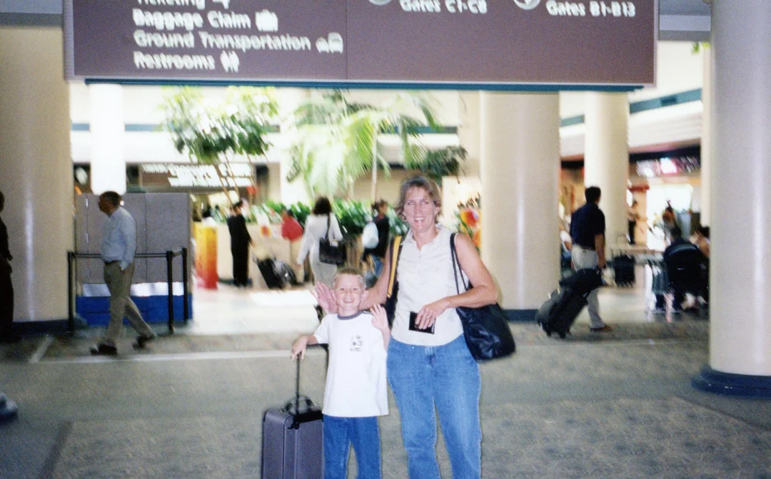 A woman and a boy with luggage stand smiling in an airport terminal under signs for gates and baggage claim. Other travelers are visible in the background.