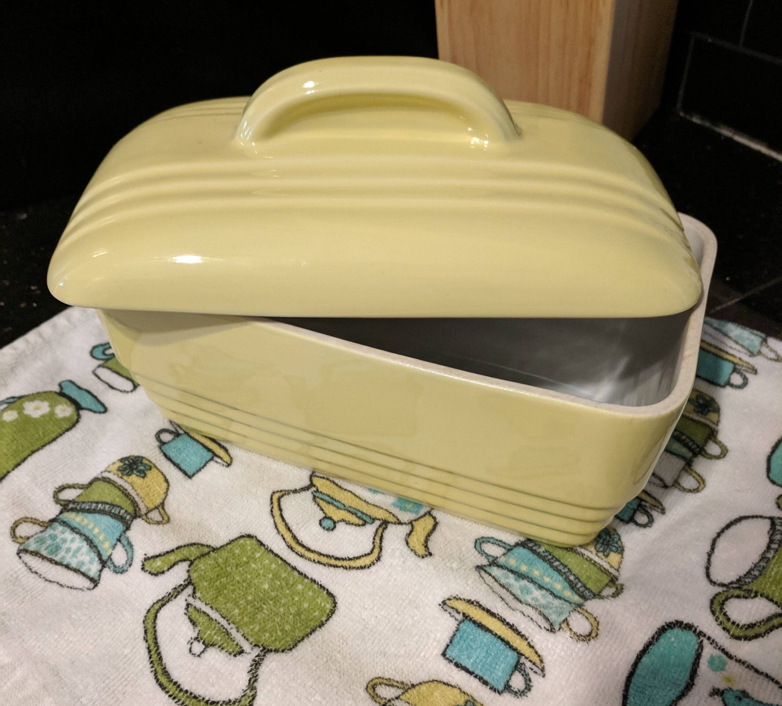 A yellow ceramic butter dish with a lid sits partly open on a kitchen towel decorated with teapots and cups.