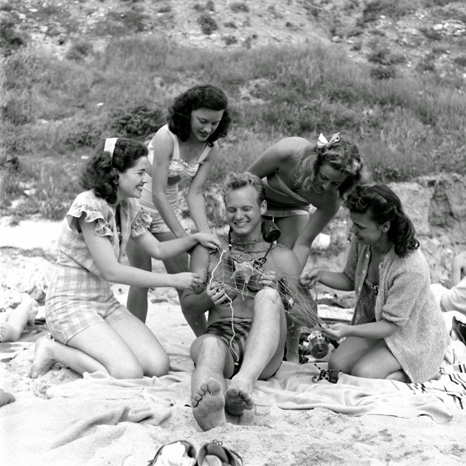 Five young women on a beach laugh as they gently cover a smiling seated man with seaweed. The group appears relaxed and playful, with grassy dunes and hills in the background.