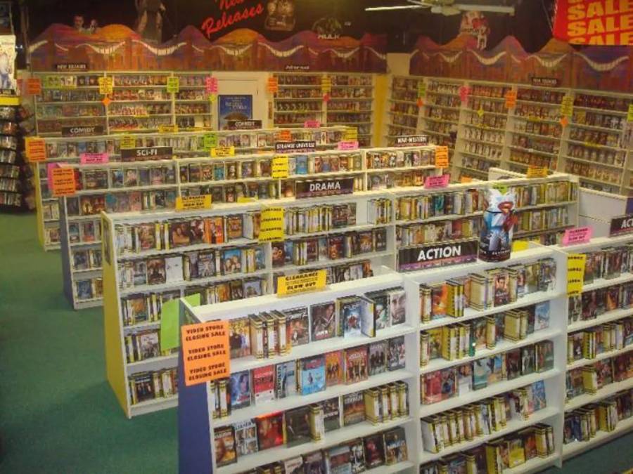 A brightly lit video rental store with multiple aisles filled with DVDs and VHS tapes, organized by genres like Sci-Fi, Drama, and Action. Colorful signs label each section.