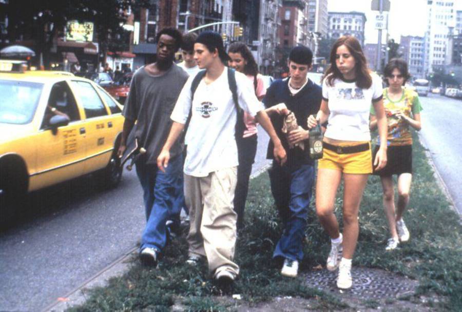 A group of teenagers walk together on a city sidewalk near a yellow taxi, with buildings and traffic visible in the background.