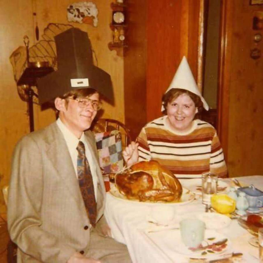 Two people sit at a table with a roast turkey. One wears a black pilgrim hat, the other a white paper hat. Both are smiling in a cozy, wood-paneled dining room.