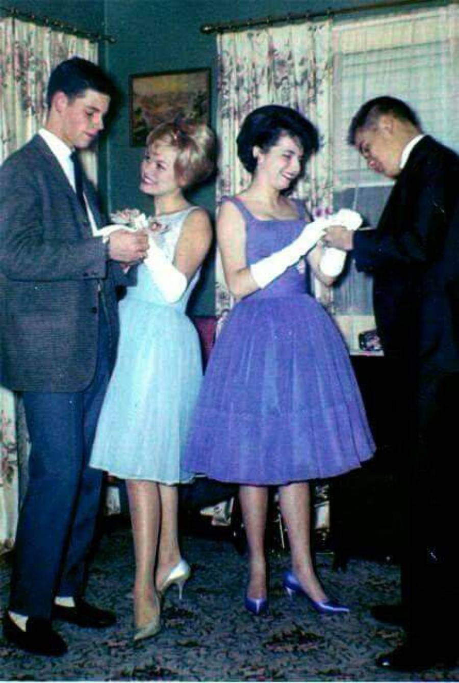 Two young couples dressed formally, with the women in pastel dresses and white gloves, smile and hold hands with their dates in a cozy, decorated living room.