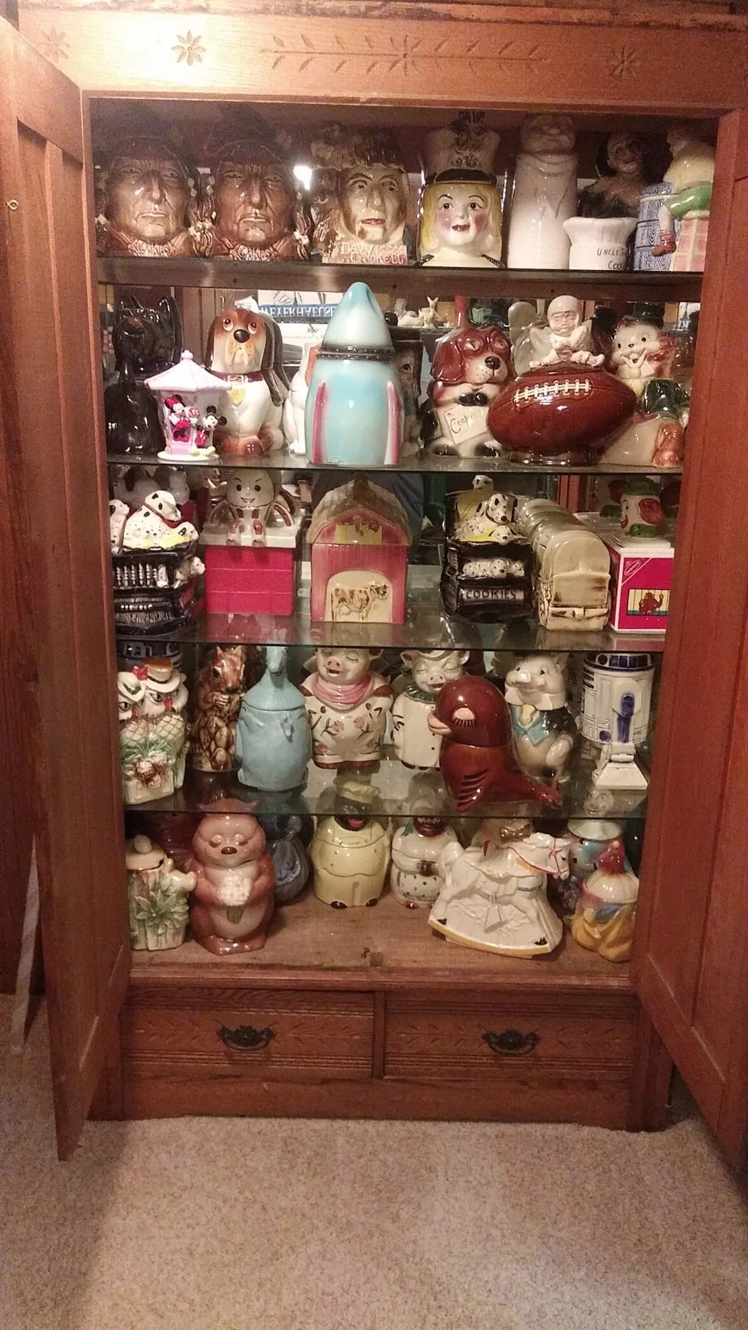 A wooden cabinet displays a large collection of colorful, assorted ceramic cookie jars shaped like animals, people, and objects, arranged neatly on glass shelves.