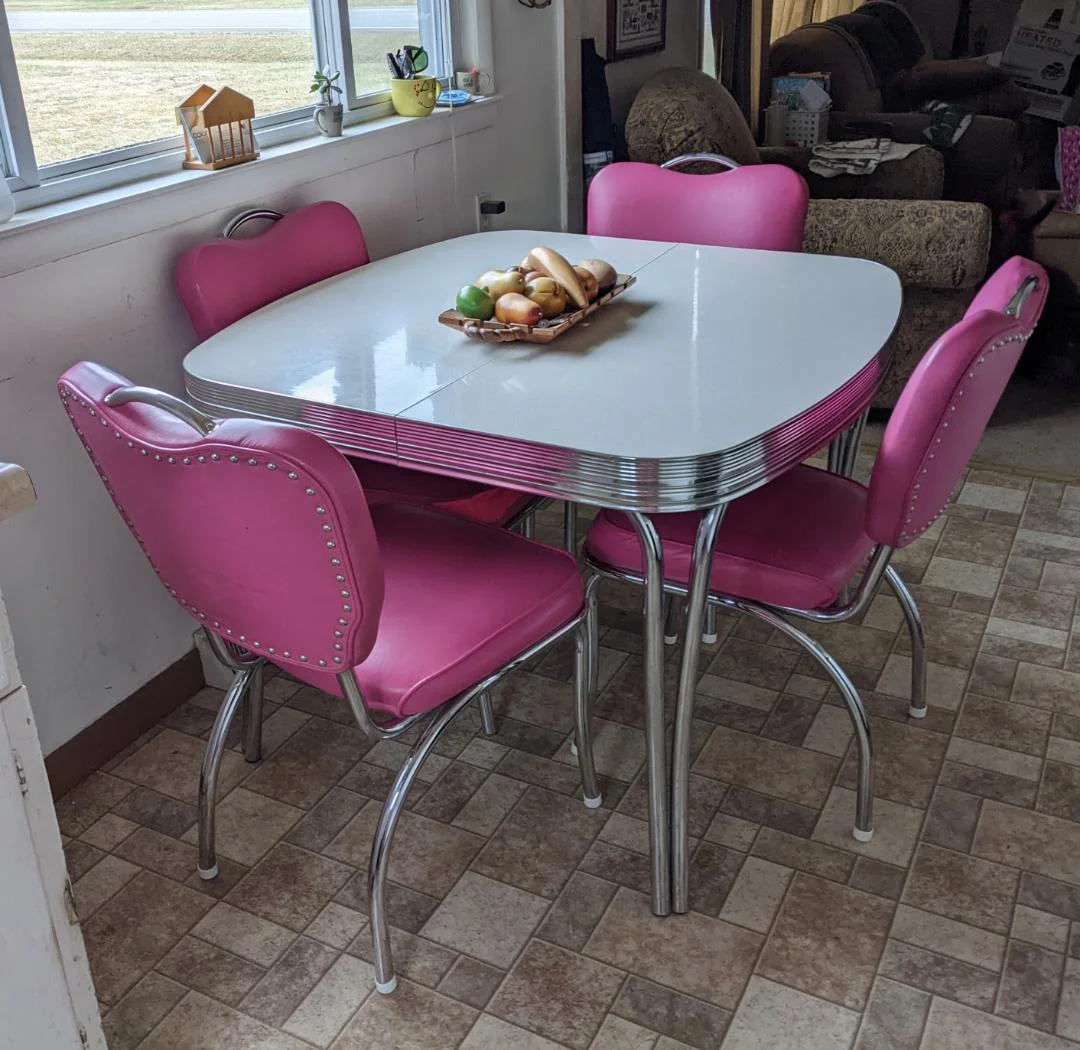 A retro dining table with a white top and chrome edges is surrounded by four pink, studded chairs. A wooden tray with fruit sits in the center. The scene is set in a room with tiled floors and sunlight from a window.