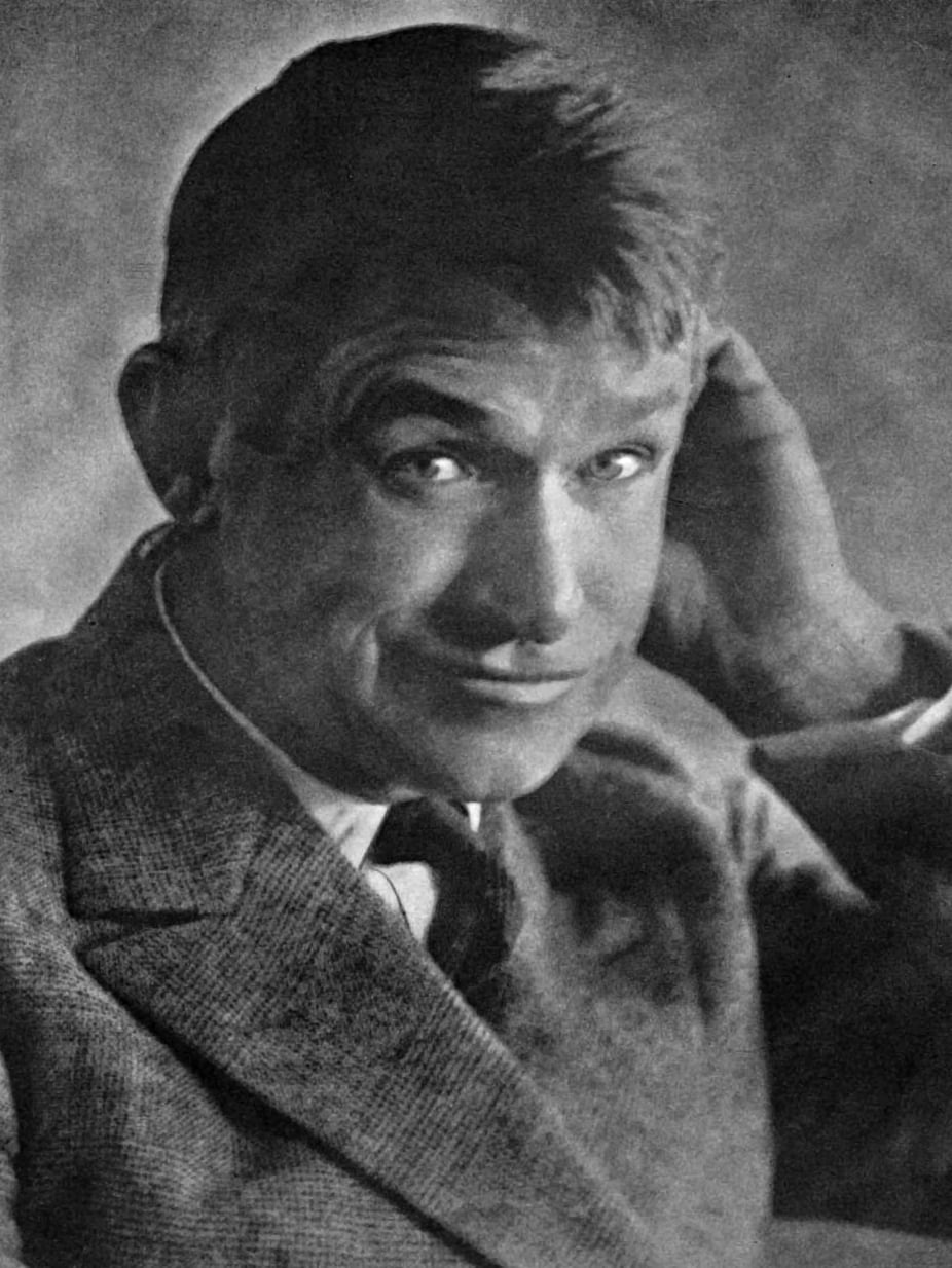 A man in a suit and tie looks at the camera, smiling slightly, with one hand behind his head. The black and white photo has a soft, vintage effect.