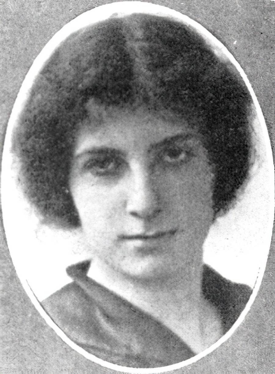 Black and white portrait of a woman with short, dark, wavy hair, looking directly at the camera. She is framed by an oval border and wears a plain, dark top. The background is light and plain.