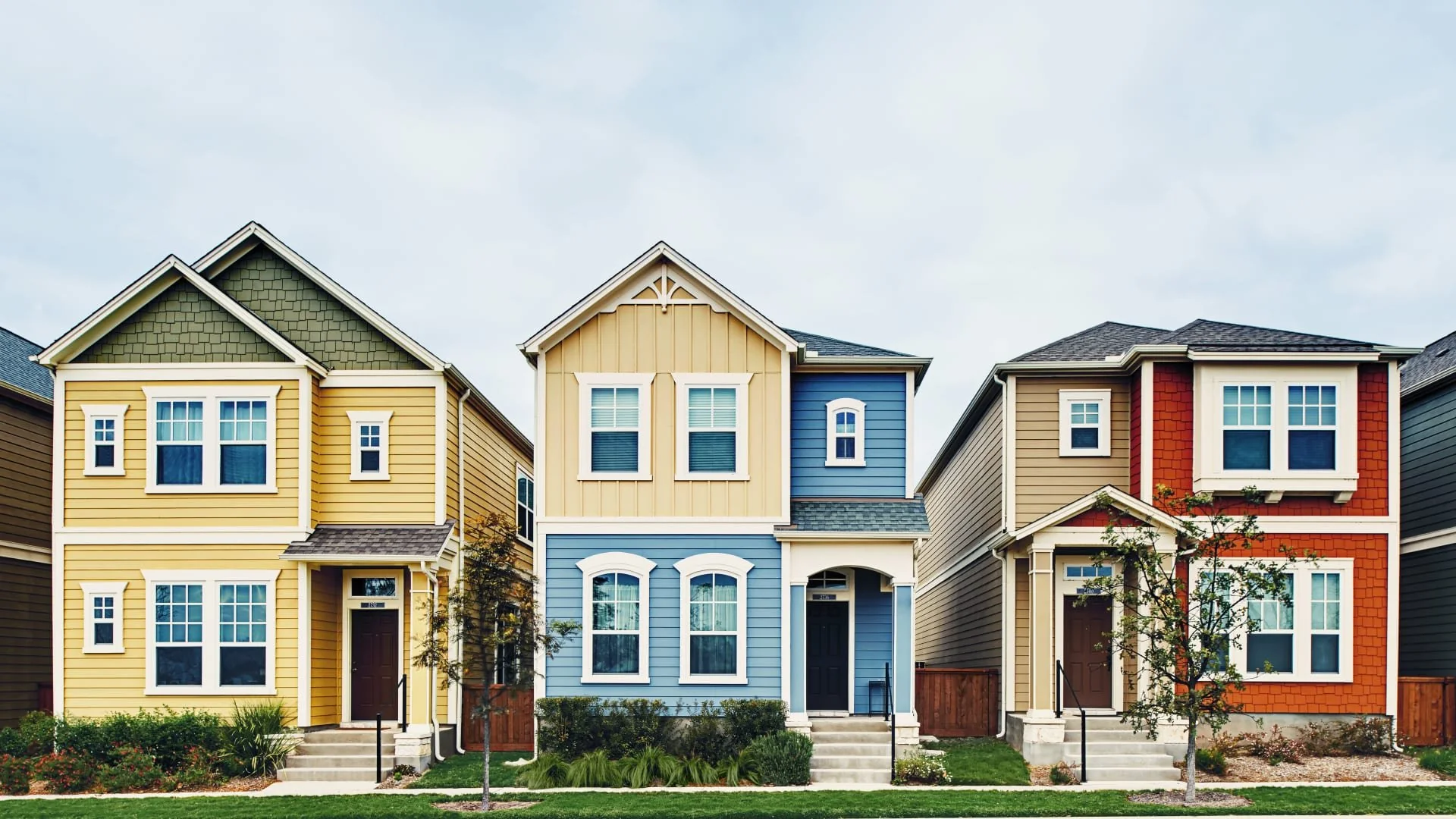 Three colorful suburban houses—yellow, blue, and red—stand side by side. Each has a small front yard, white trim, and a sidewalk in front. The sky is overcast, and there are young trees and shrubs in the yards.