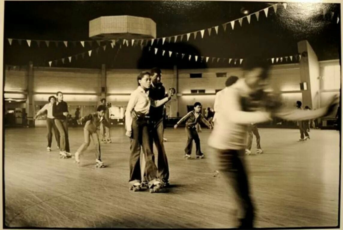 Black-and-white photo of people roller skating indoors under hanging flags. Some skaters are in focus while others are blurred from motion, creating a lively scene.