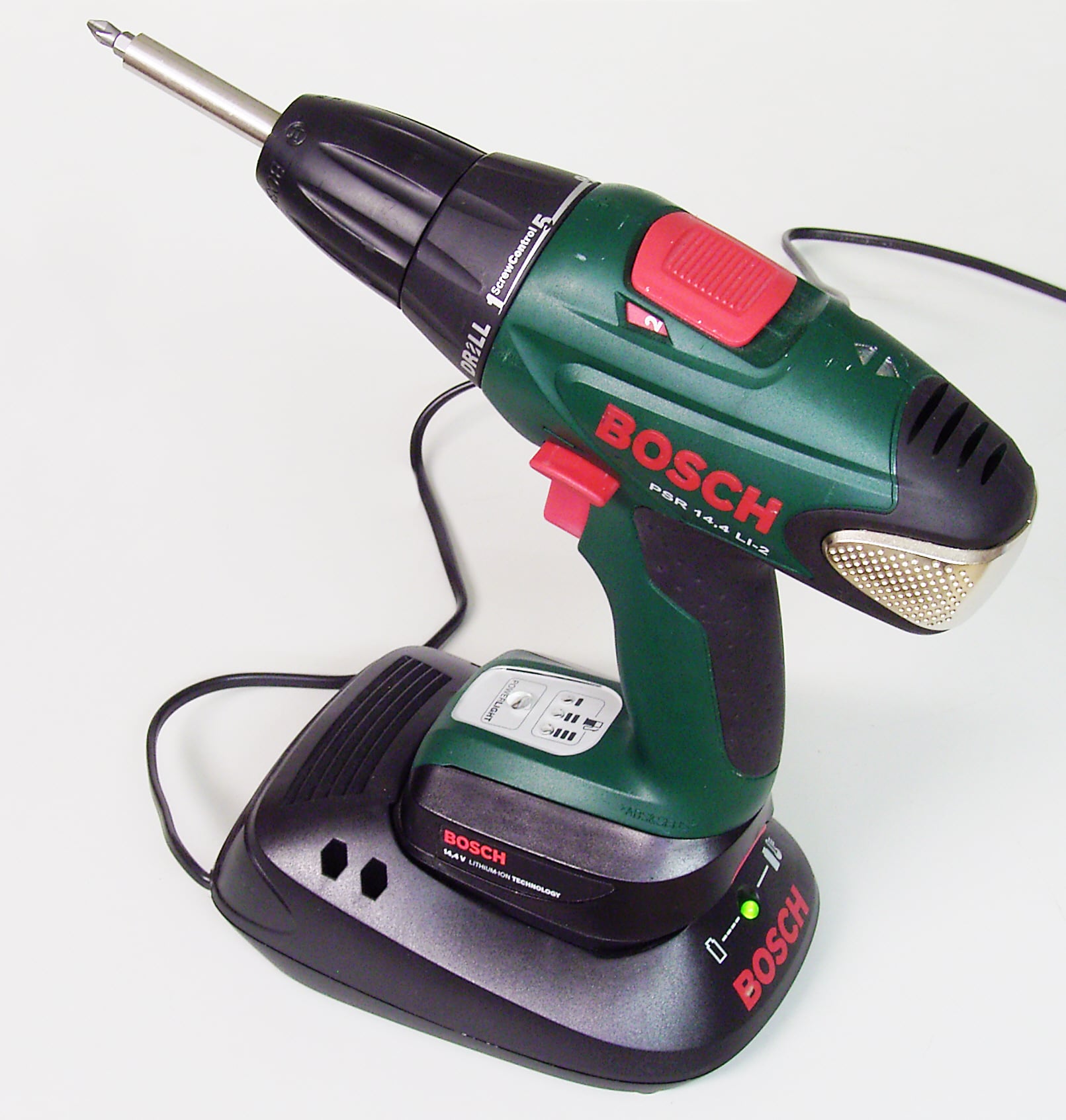 A green Bosch cordless power drill is placed on its black charging station, with a power cord attached. The drill has red buttons and branding, and a metal drill bit is installed.