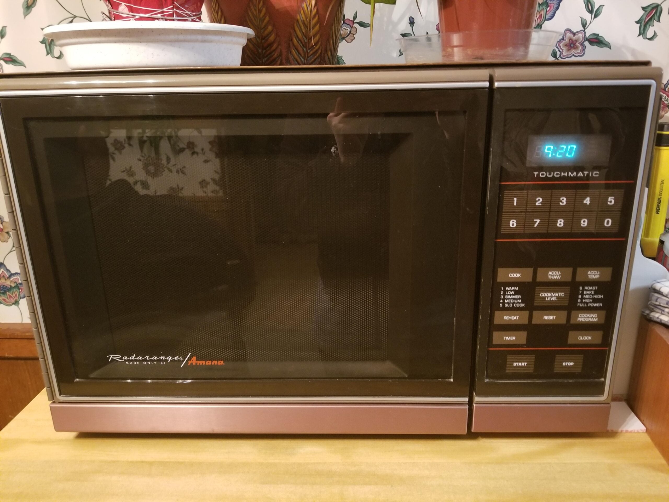 A vintage Amana Radarange microwave with a digital keypad and display showing 9:20, placed on a yellow countertop with floral wallpaper and potted plants in the background.