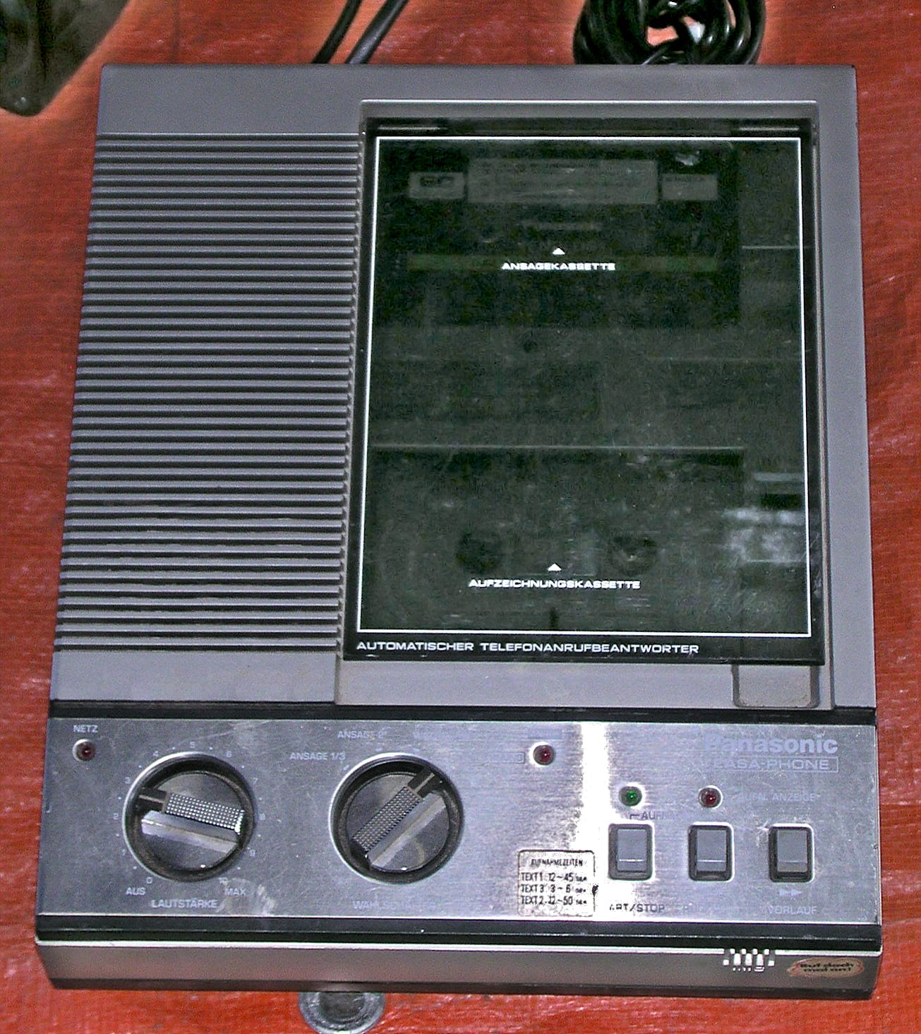 A vintage Panasonic telephone answering machine with buttons, dials, and a cassette slot, placed on a red surface with a visible power cord.
