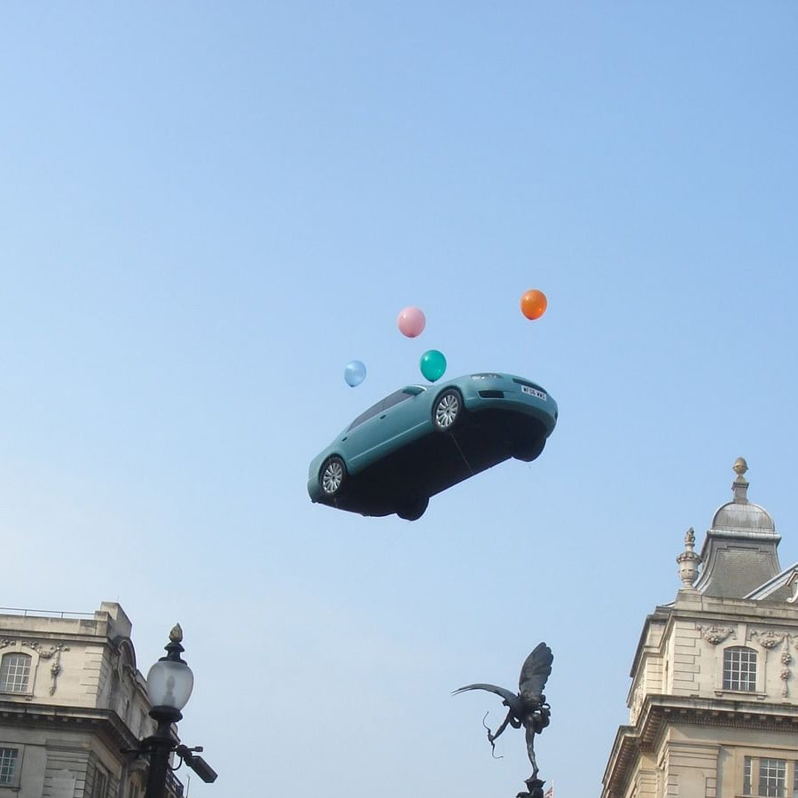 A teal car-shaped balloon floats in the sky above buildings, surrounded by colorful balloons, with a statue and lamppost below.