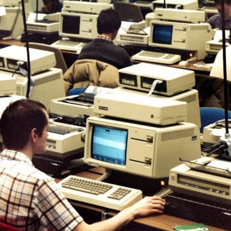 Several people work at vintage Apple computers in a crowded computer lab, with beige monitors, printers, and keyboards filling the desks. The setting appears to be from the 1980s.