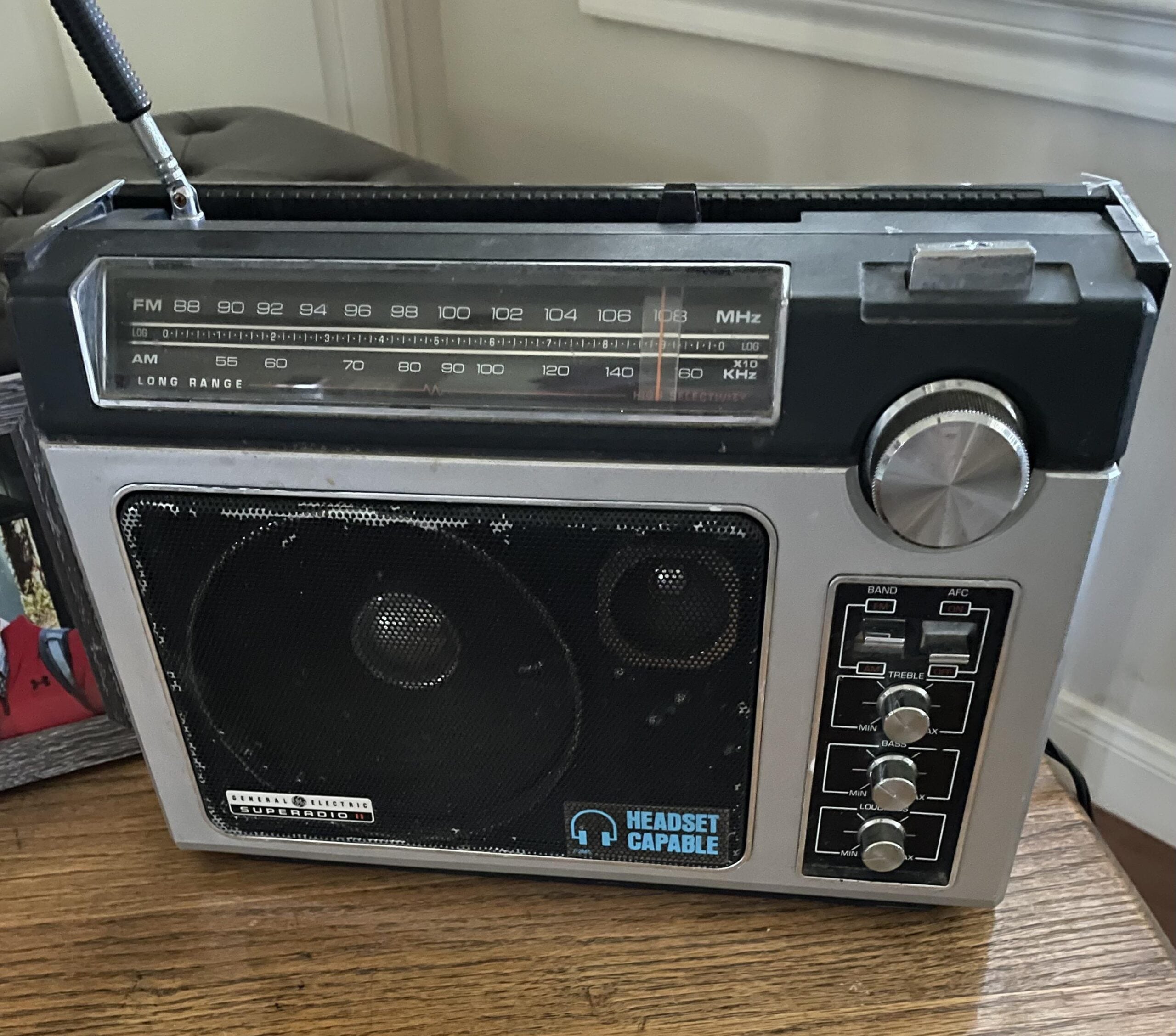A vintage portable radio with an antenna, large tuning dial, speaker, and buttons, sitting on a wooden surface near a window. The radio has an AM/FM display and a “headset capable” label.