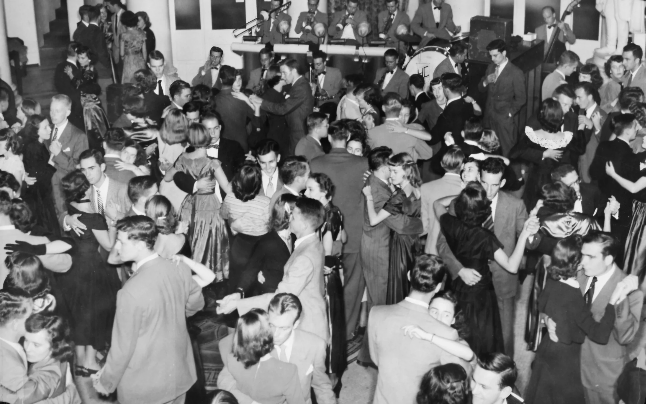 Black and white photo of many couples dressed formally, dancing closely at a crowded event or party. A live band is performing on a stage in the background.