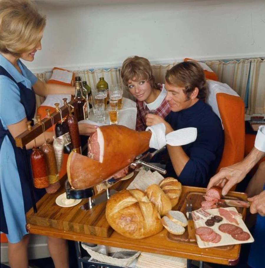 Two airline passengers are served meats, bread, and drinks by flight attendants in vintage uniforms onboard a plane. A large ham and various deli meats are displayed on a cart.