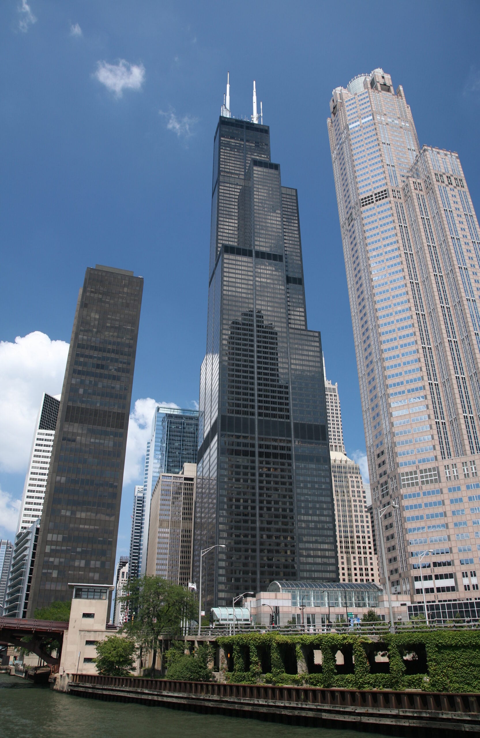 Tall black skyscraper with antennae rises between two other high-rise buildings against a blue sky, with a river and green trees in the foreground.
