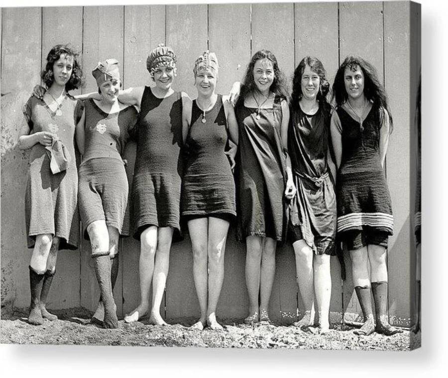 Seven women in vintage swimsuits and swim caps stand barefoot in a row, smiling, with arms around each other in front of a wooden wall. The image appears to be from the early 20th century.