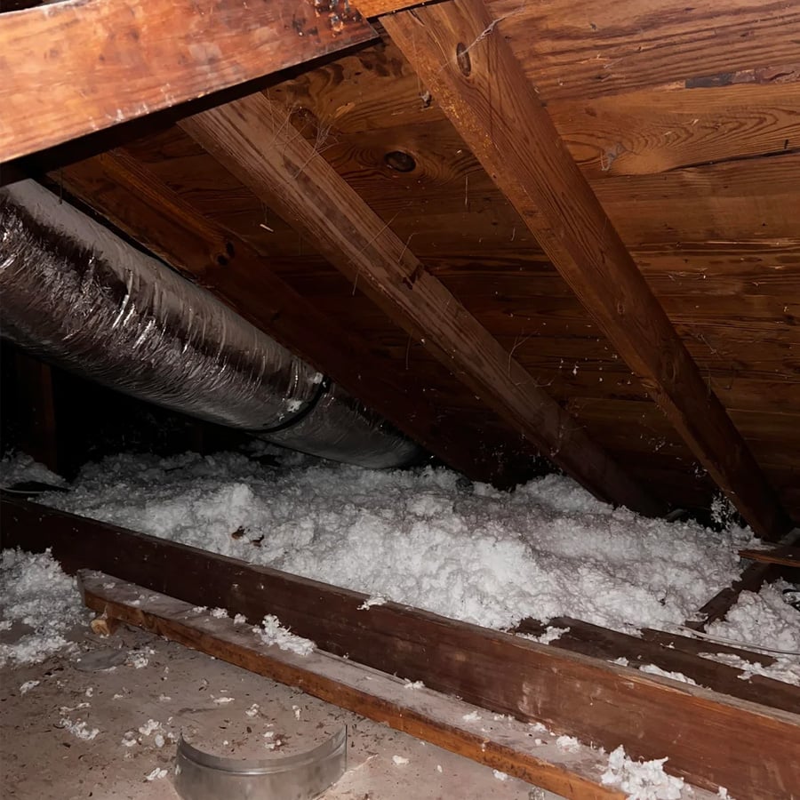 Attic with exposed wooden beams, ductwork, and loose white insulation covering the floor, dimly lit.