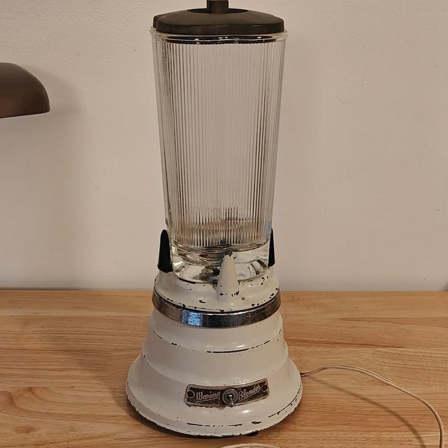 A vintage white blender with a ribbed glass jar sits on a wooden surface against a plain wall. The base shows signs of wear and has a visible power cord.
