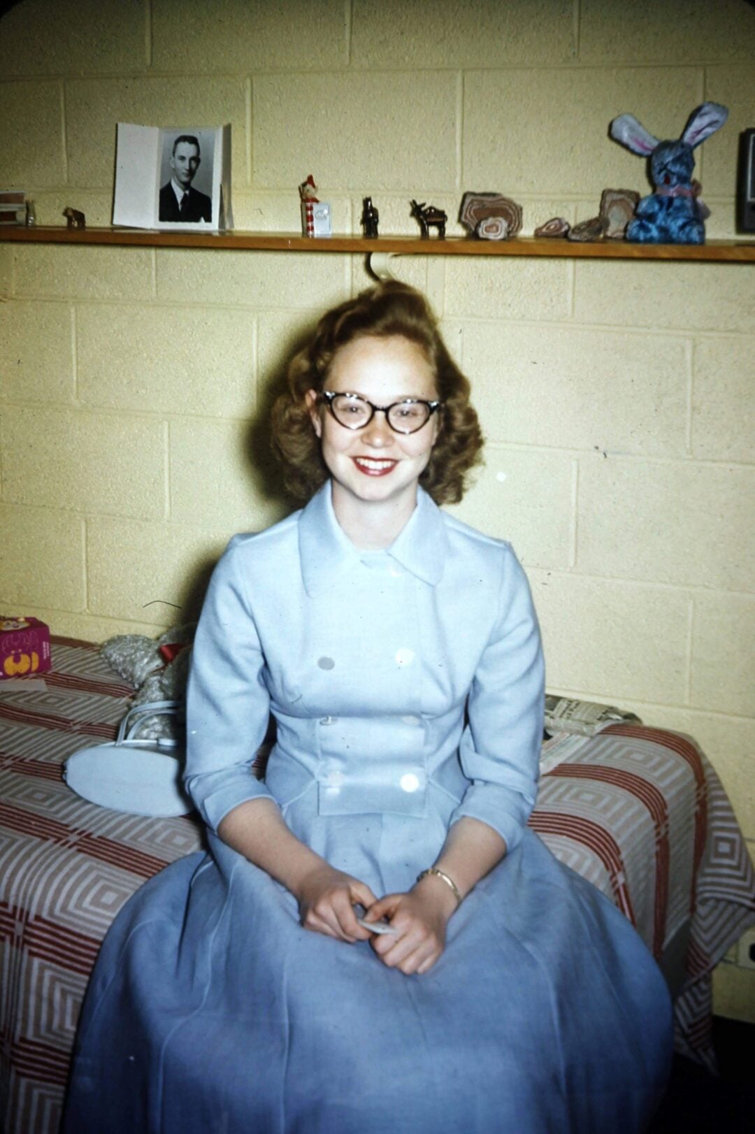 A young woman with curly hair and glasses, wearing a light blue dress, sits smiling on a striped bed in a room with a shelf holding photos, figurines, and a blue stuffed animal behind her.