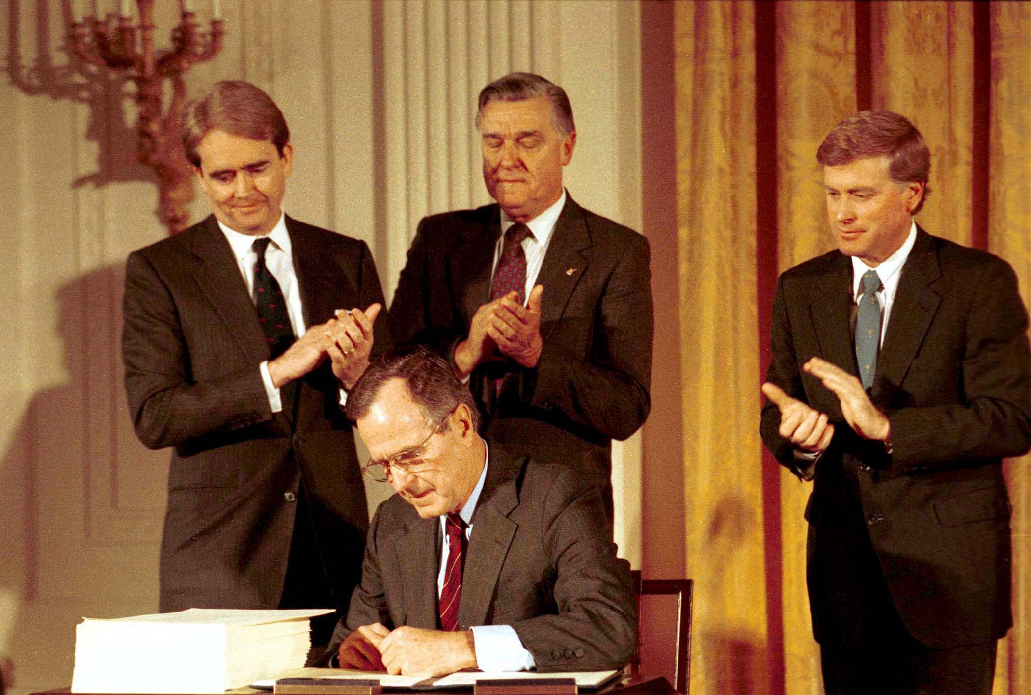 A man in a suit sits at a desk signing papers while three men standing behind him clap. The setting appears formal with elegant decor in the background.