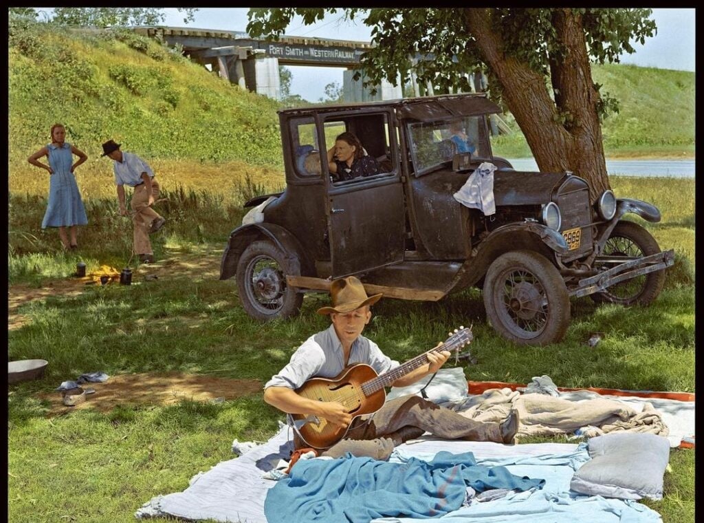 A man plays guitar on a blanket near an old car under a tree; two people stand by the car and a woman stands apart, with a bridge and grass in the background.