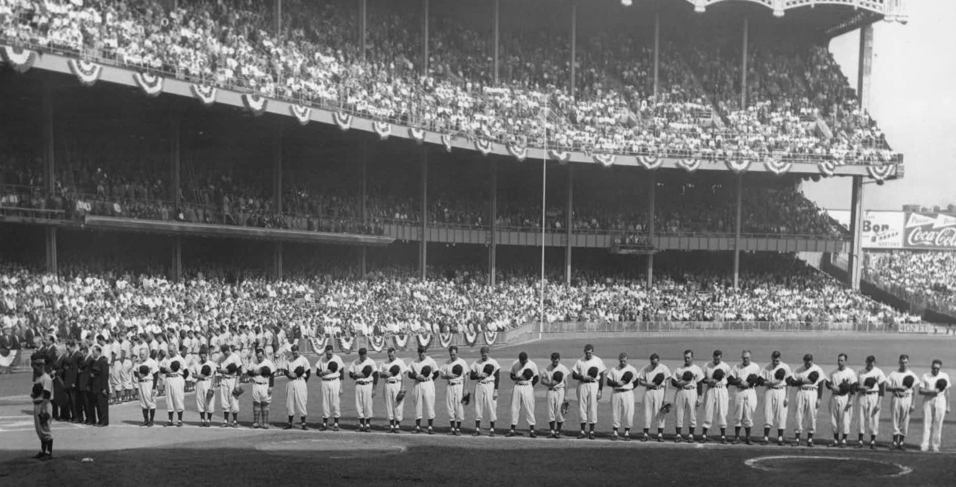 Baseball players line up on the field for the national anthem, with hands on their hearts. The stadium is packed with fans and decorated with bunting.