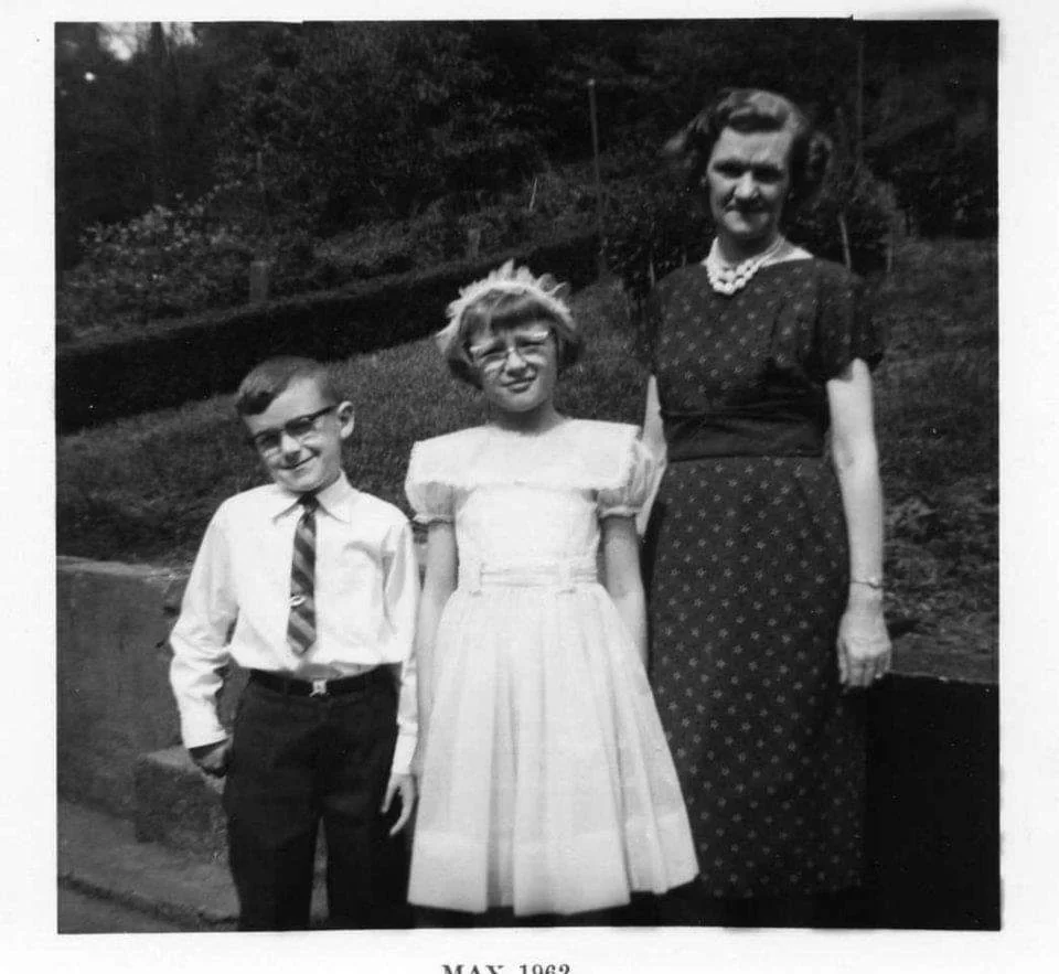A boy in a tie, a girl in a white dress, and a woman in a dark dress stand outdoors, smiling, likely posing for a special occasion. The photo is black and white and dated May 1962.