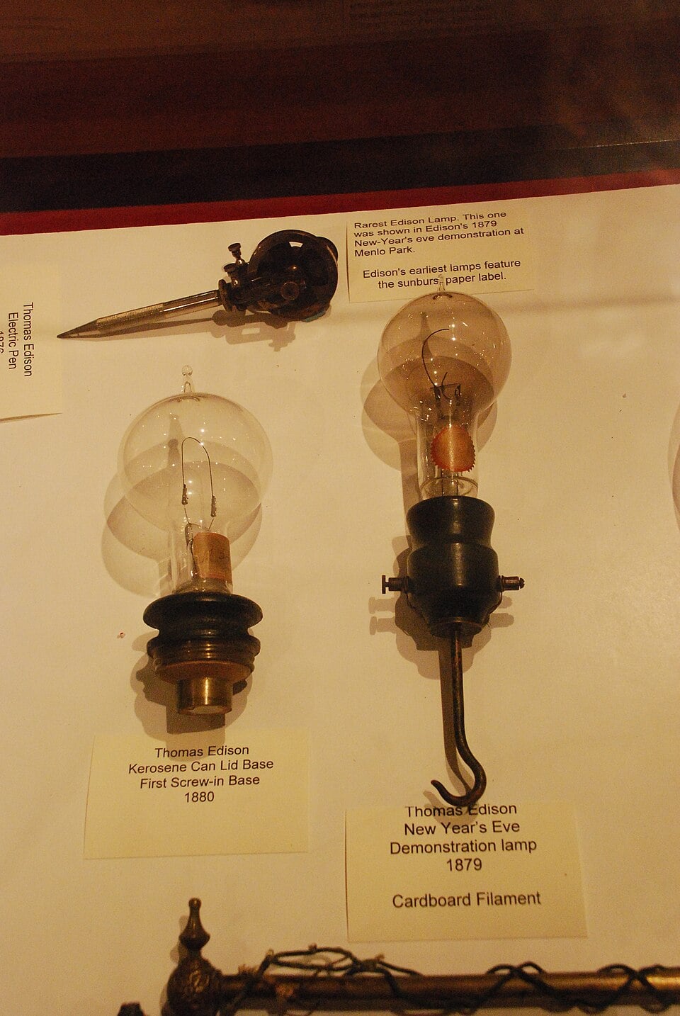 Two vintage Edison light bulbs with descriptive labels sit on display, showing early electric lamp designs from 1879 and 1880, each with distinct bases and filaments, on a white surface with additional historical lighting items nearby.