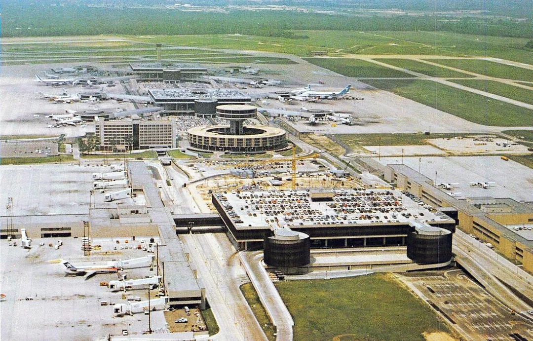 Aerial view of a large airport with multiple terminals, airplanes on the tarmac, parking lots full of cars, and runways surrounded by green fields.