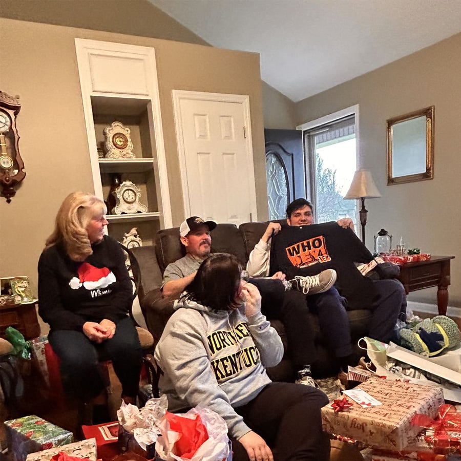 Four people sit on sofas in a living room filled with wrapped presents. One person holds a “WHO DEY” sign. The atmosphere is festive, with holiday decorations and a Christmas hat visible.