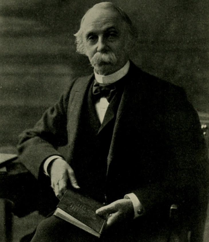 An older man with a mustache and beard sits holding a closed book, wearing a dark suit, bow tie, and vest, looking toward the camera. The background is plain and softly lit.