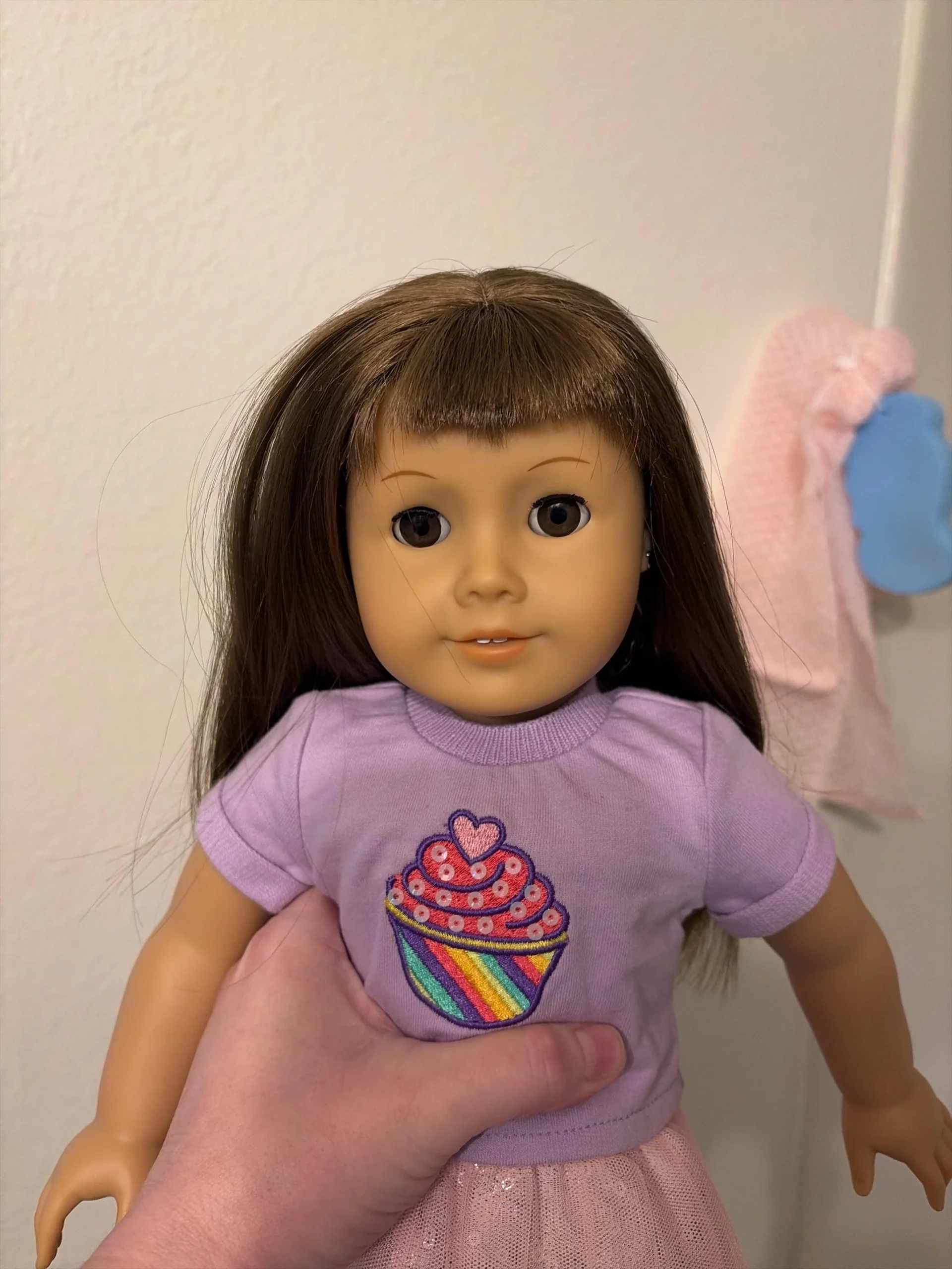 A hand holds a doll with long brown hair, brown eyes, and a lavender shirt featuring a colorful cupcake graphic. There is a light pink cloth and blue shape hanging in the background.