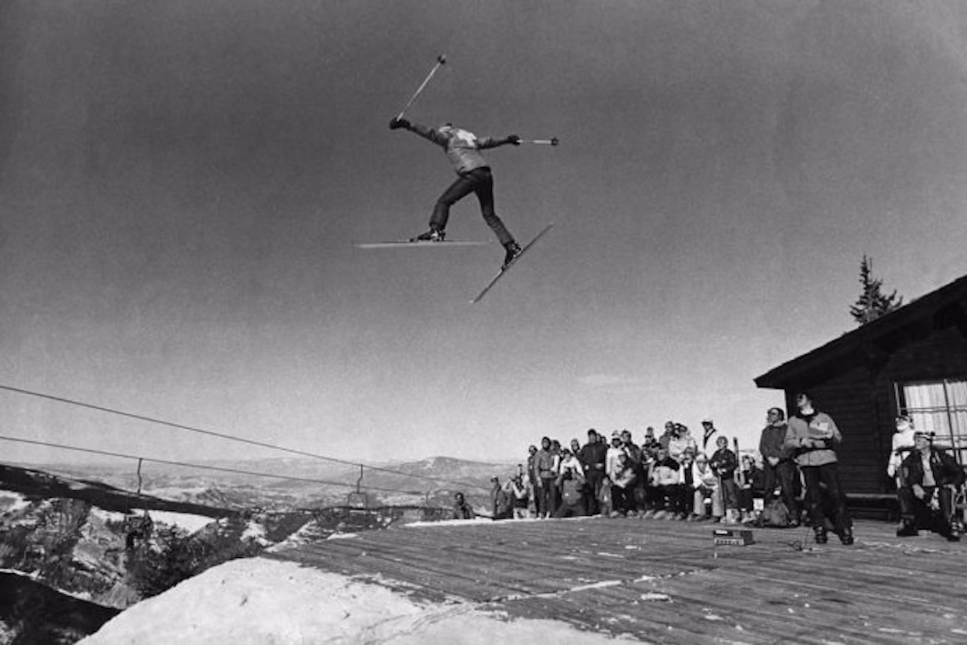 A skier in mid-air performs a jump over a wooden platform, watched by a group of people standing near a cabin with snowy mountains in the background.