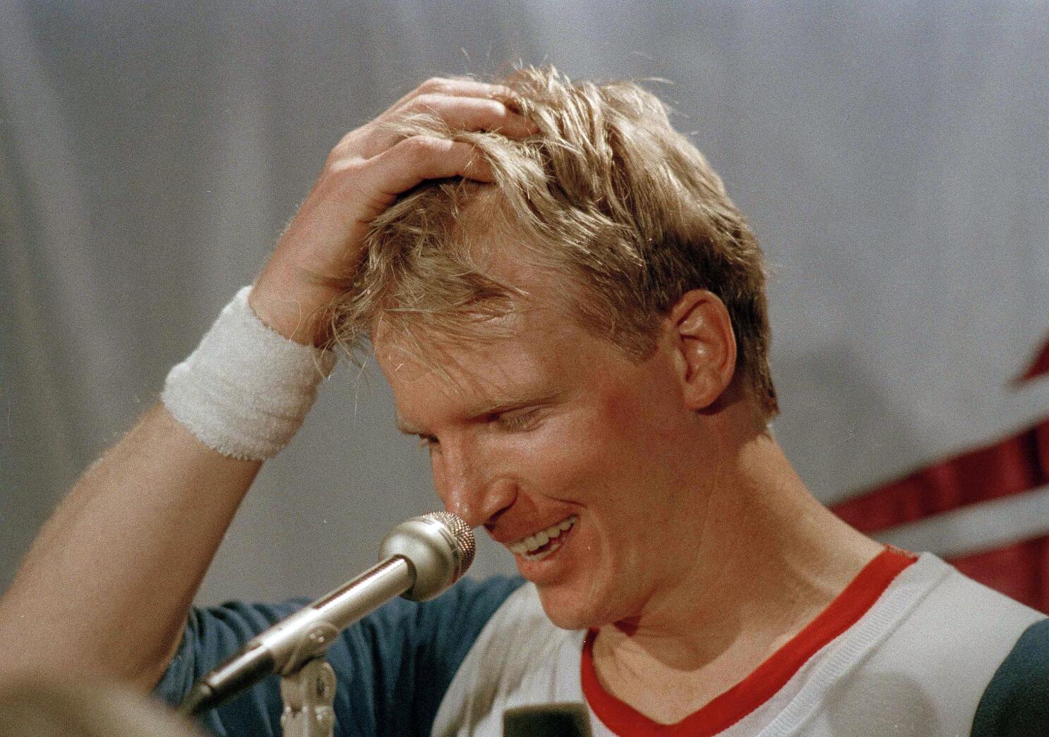 A smiling athlete with short blond hair and a white wristband scratches his head while speaking into a microphone.