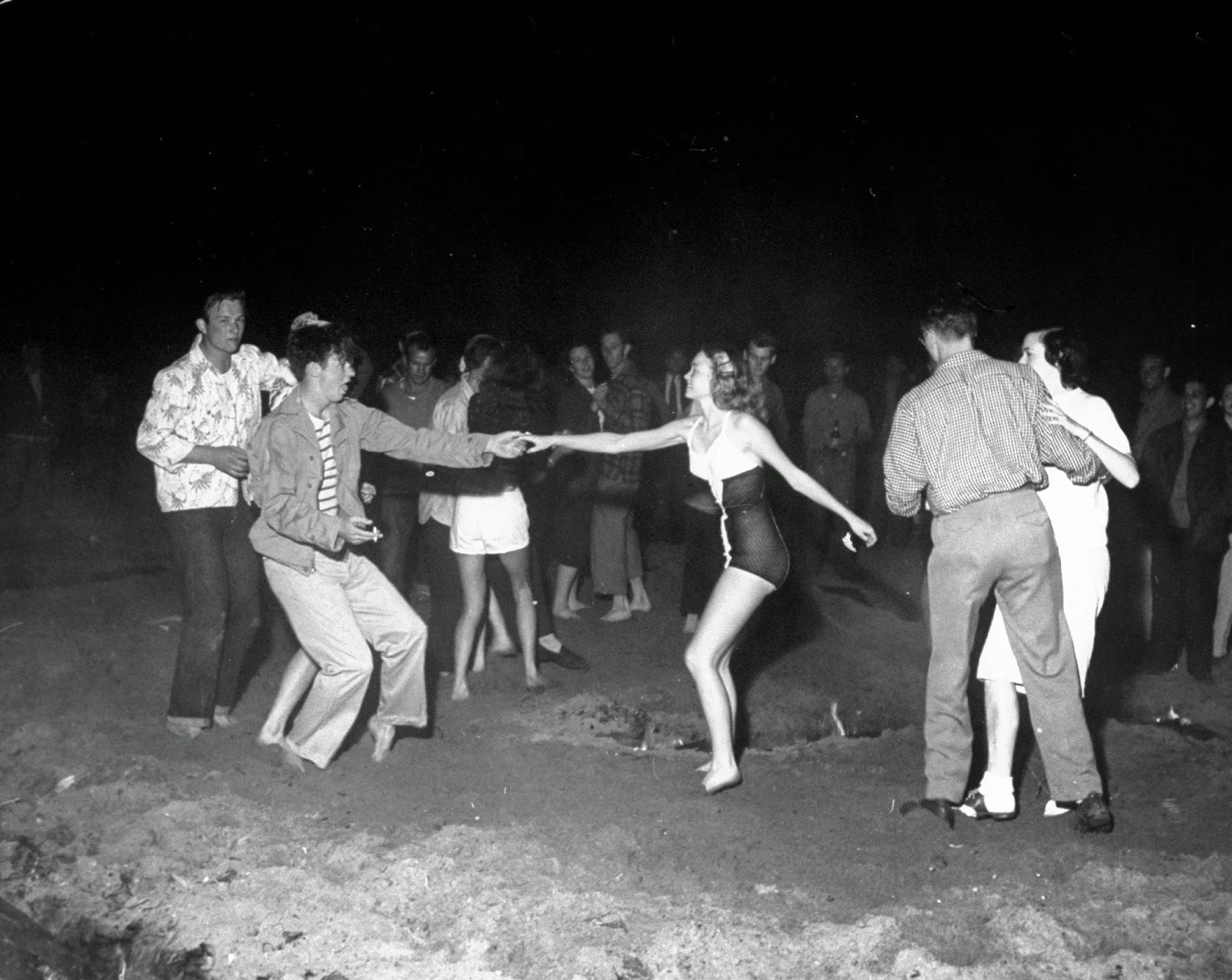 A group of people dance barefoot outdoors at night, forming couples. Most are dressed in casual 1960s clothing, while one woman wears a swimsuit. Others watch in the background. The scene is lively and joyful.