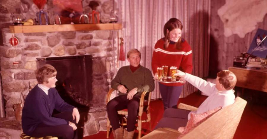 Four people relax in a cozy living room with a stone fireplace. One woman serves drinks to three seated people. The room has red carpet, wood paneling, and vintage decor.