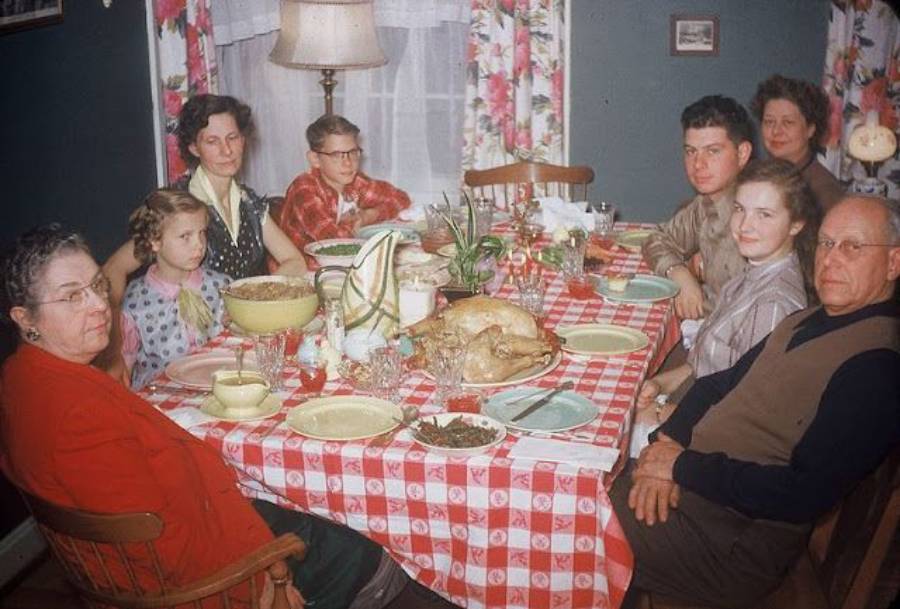 Eight people sit around a table with a red checked tablecloth, sharing a meal including a roast turkey. The room has floral curtains and a lamp in the background.