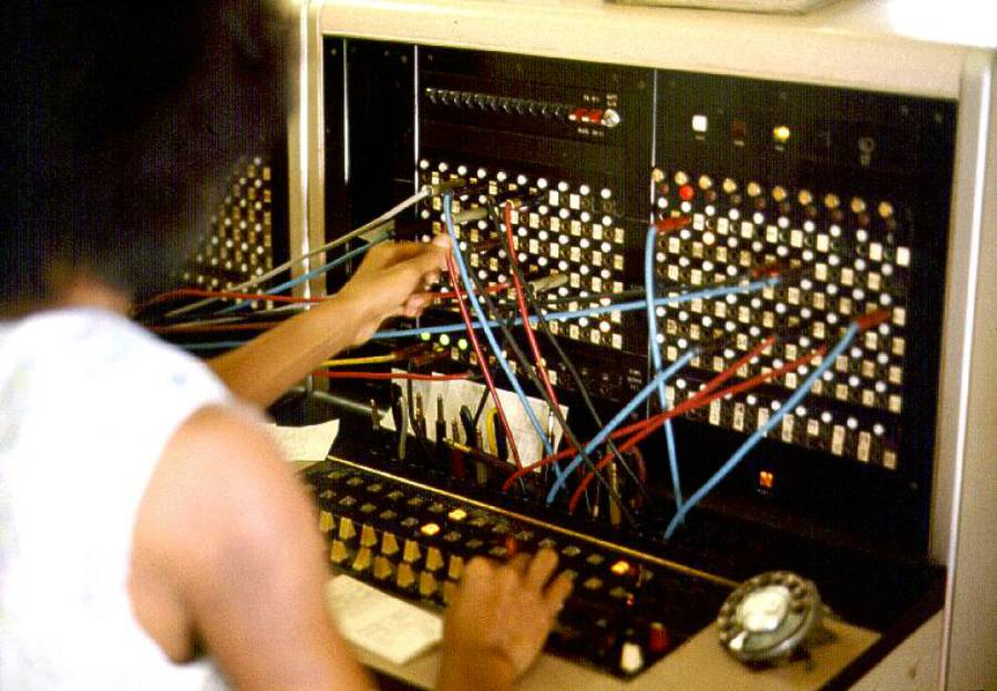 A person operates a vintage telephone switchboard, plugging in cables and pressing buttons to connect calls. A rotary dial phone sits on the counter nearby.