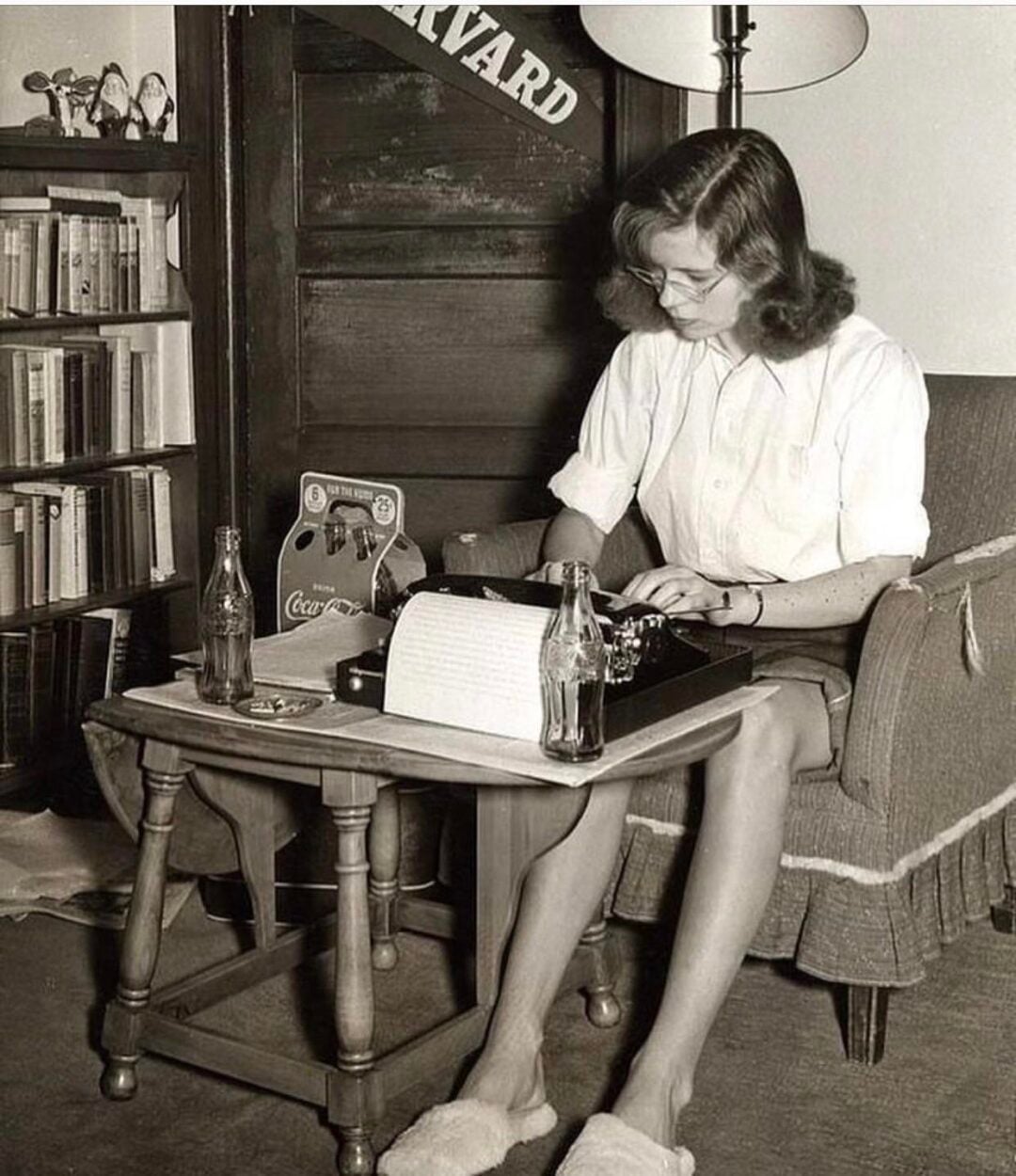 A woman in glasses and slippers sits at a small table, typing on a typewriter. Two glass Coke bottles and books are nearby. A “Harvard” banner hangs behind her. The setting appears vintage and cozy.