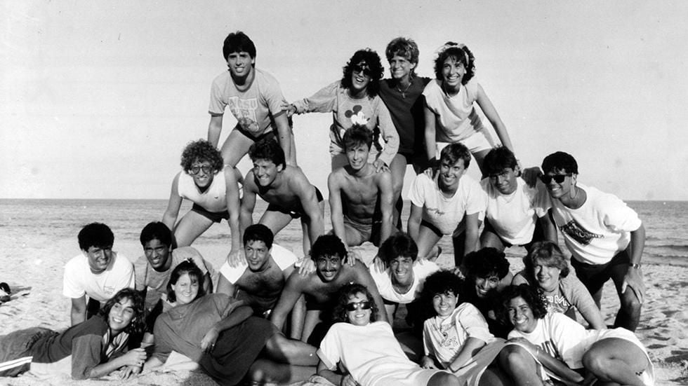 A large group of young people form a human pyramid on a sandy beach, smiling and posing for the camera with the ocean in the background.