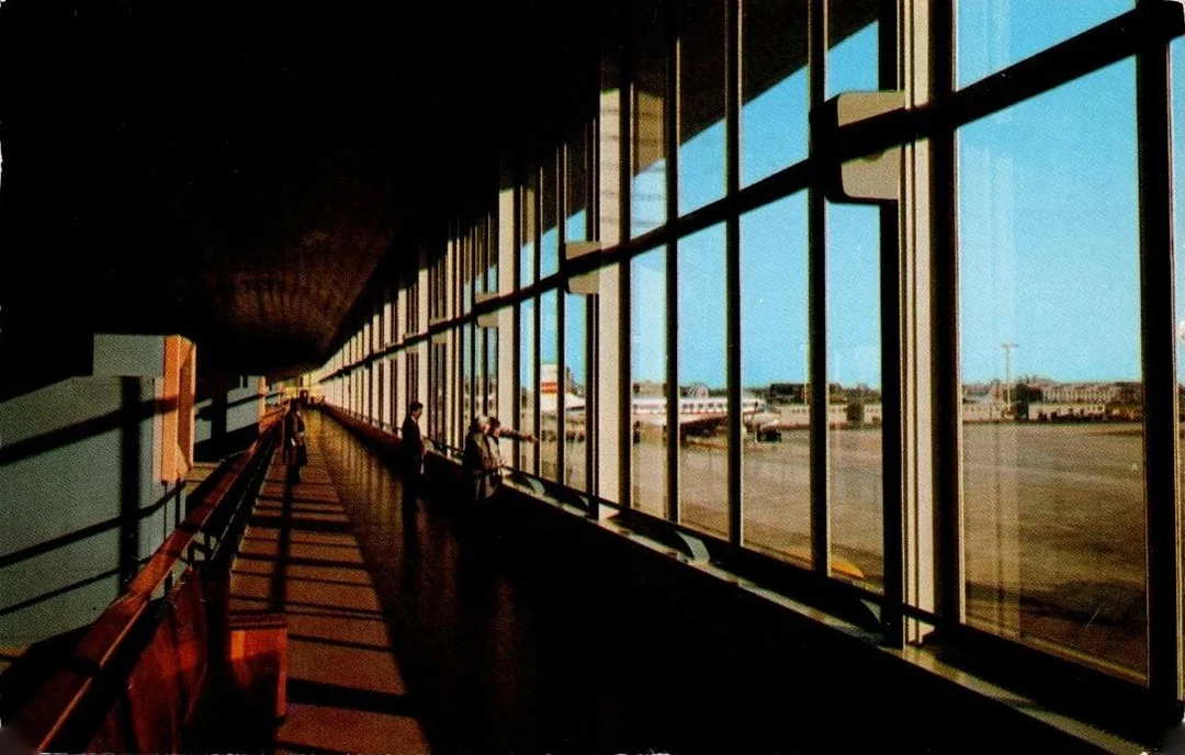 Long corridor with large glass windows overlooks an airport tarmac in daylight. Few people are visible inside, and planes are seen outside. Shadows stretch across the floor, creating a pattern.
